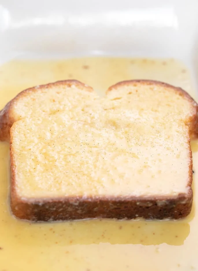 Slice of brioche bread soaking in egg mixture in white casserole dish