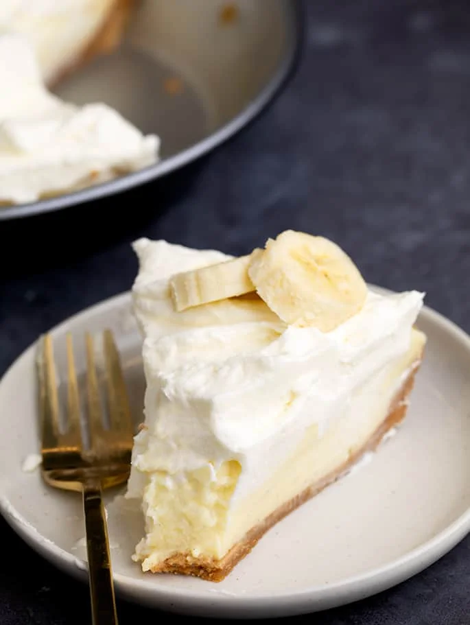 Slice of pie with brown graham cracker crust, yellow custard, whipped cream and bananas on small white plate with fork