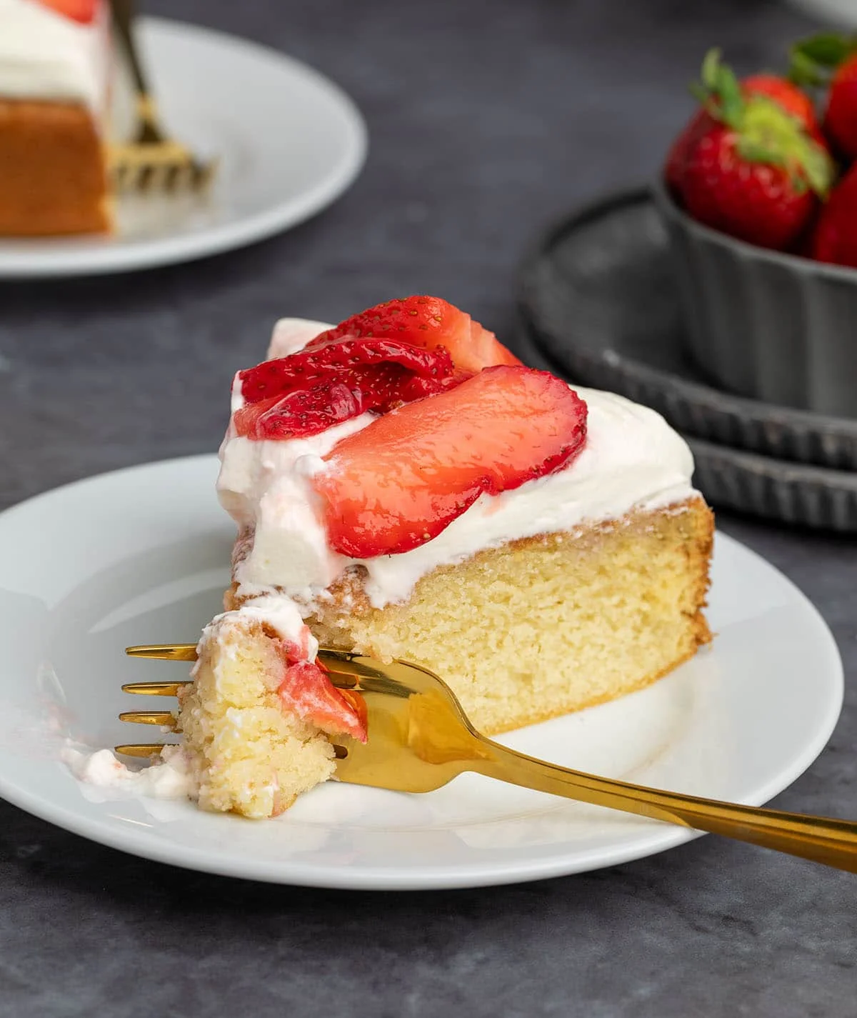 slice of yellow cake with white whipped cream and red strawberries on small white plate with gold fork pressed into tip of cake