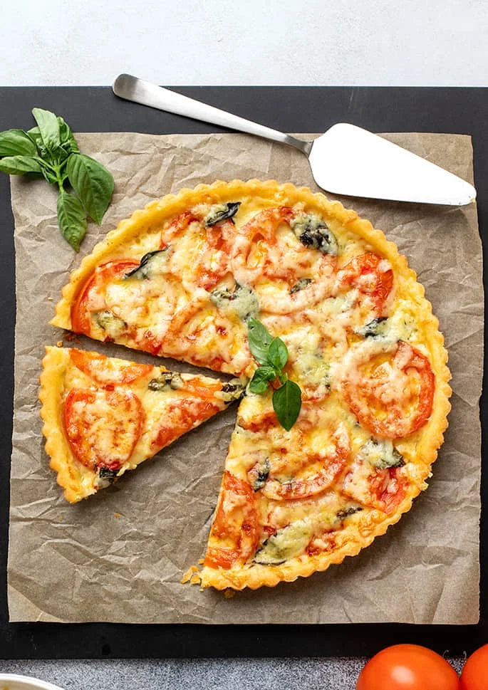 Sliced tomato tart on brown paper and a black board with pie server, basil leaves and tomatoes.