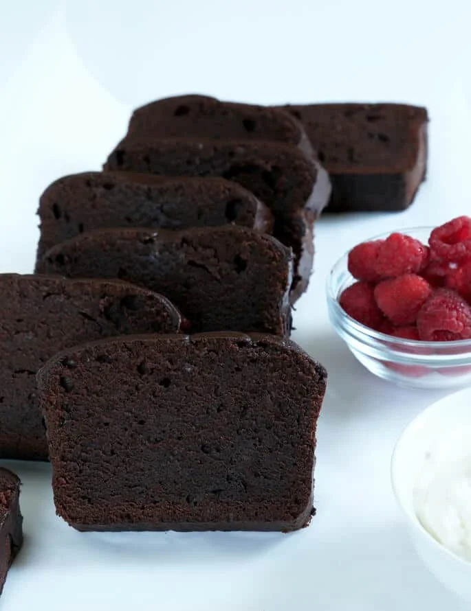 Slices of chocolate pound cake leaning against each other on a white surface with a bowl of raspberries in back