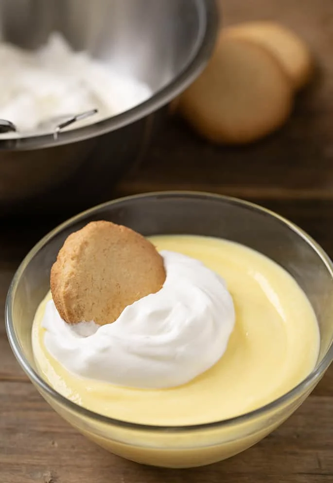Small glass bowl with yellow custard, whipped cream, vanilla wafer, more cookies in background