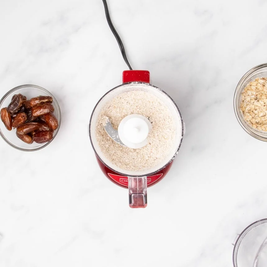 small red mini chop food processor with clear plastic bowl with ground oats, dates in a bowl on the side