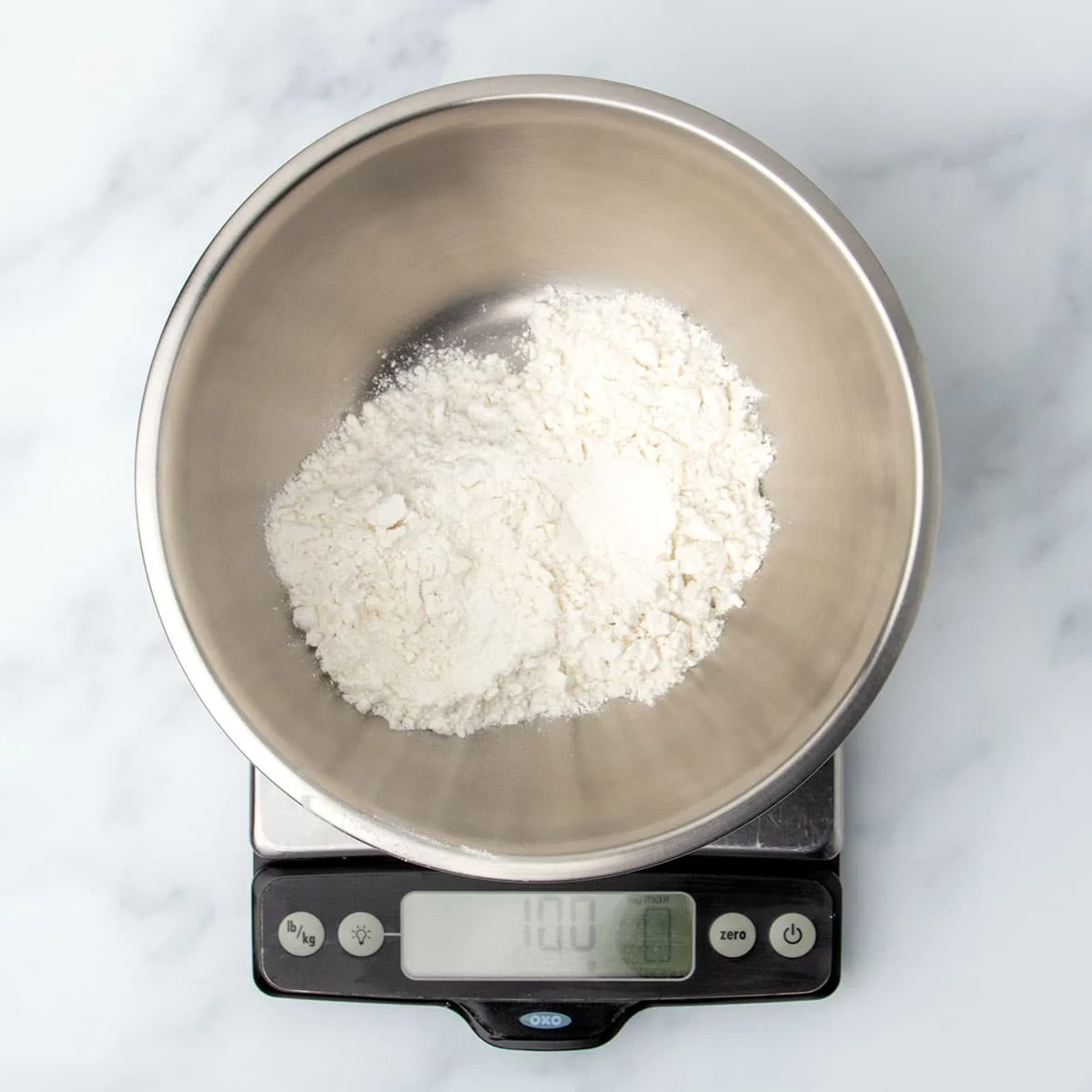 small round metal mixing bowl with white powder on a digital kitchen scale reading 100 grams