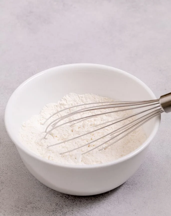 Small white bowl with flours and a large whisk.