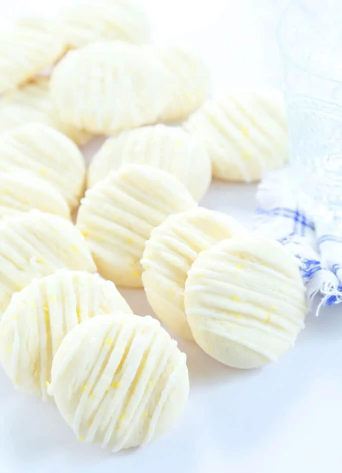 Small white cookies with yellow flecks on white surface with lemon zest on top and white and blue striped cloth