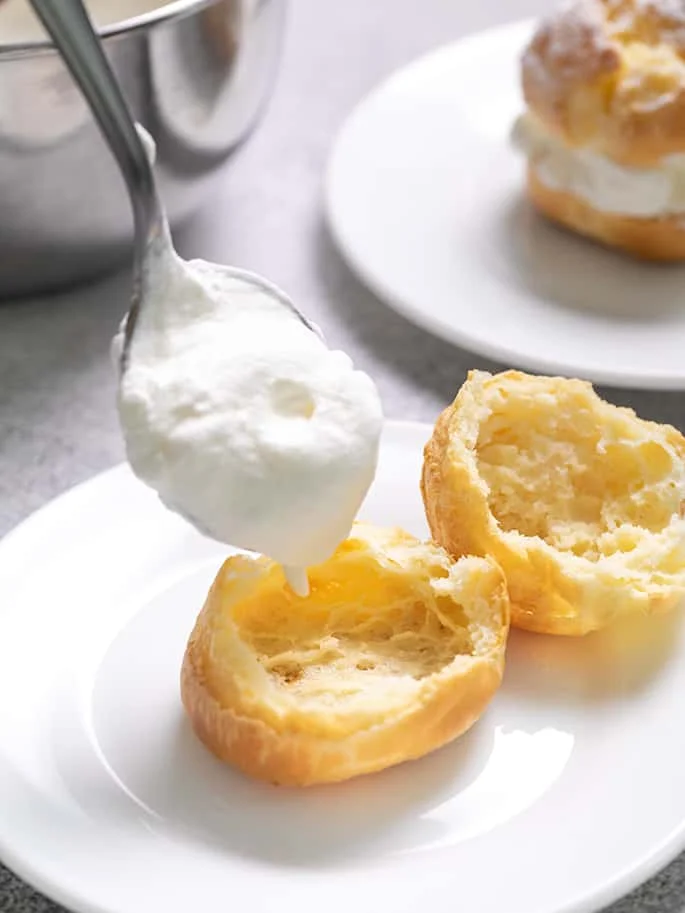 Spoon placing whipped cream into hollow half of baked choux pastry on small white plate