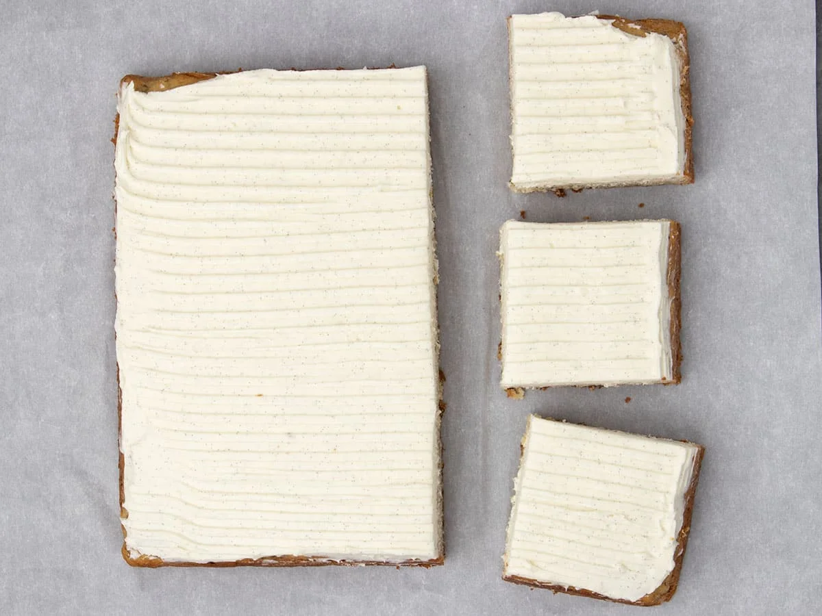 square banana cake with top covered in white cream cheese frosting with 3 squares cut, all on white paper
