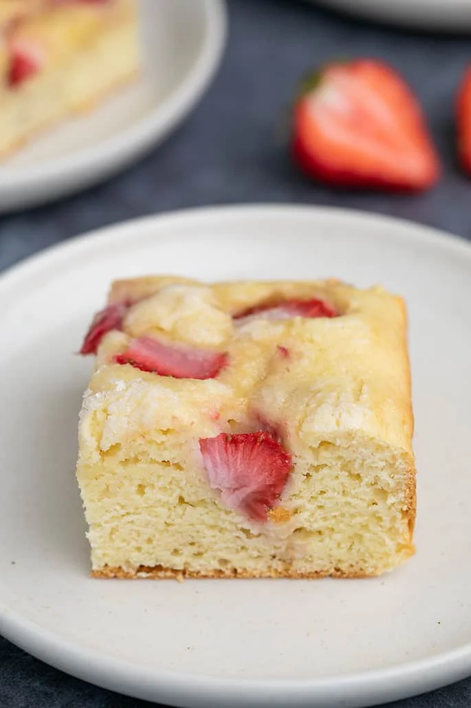 Square of strawberry gluten free dessert on small white plate with cut strawberry in background