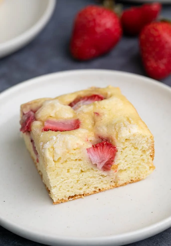 Square piece of strawberry gluten free dessert on small white plate with strawberries in background