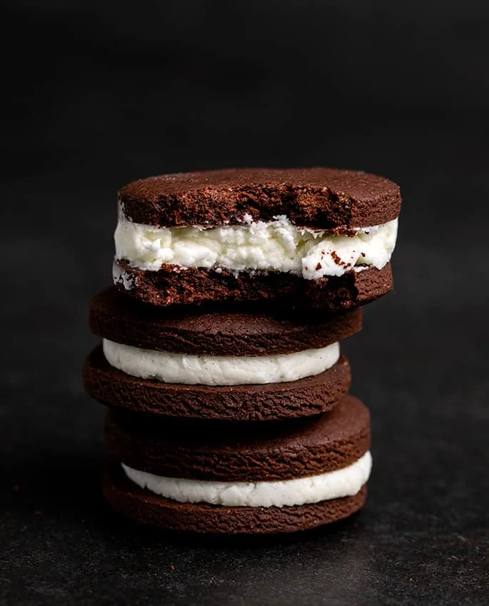 Stack of 3 chocolate sandwich cookies with white filling with a bite taken from the top cookie, all on black surface