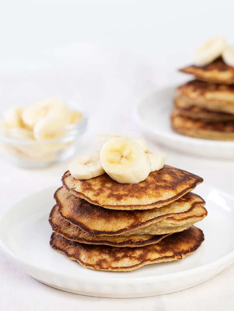 Stack of 4 banana pancakes with banana slices on top on white plate.