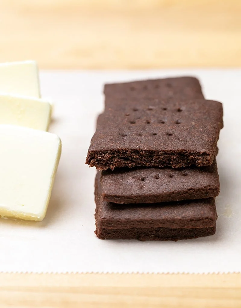 Stack of 4 chocolate rectangular cookies and pats of yellow butter on white paper on blond wood table
