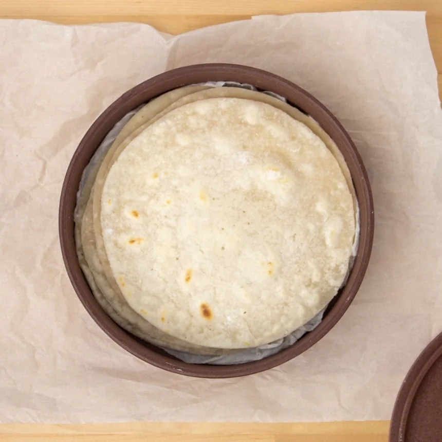 stack of cooked tortillas with some golden brown spots, all in brown round plastic tortilla warmer with wet white paper towels underneath, in the warmer