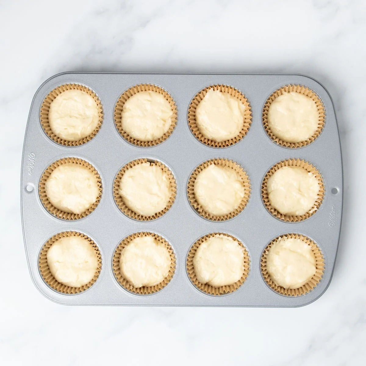 standard 12 cup metal muffin tin with brown muffin liners in wells filled 2/3 of the way full with light yellow raw cupcake batter