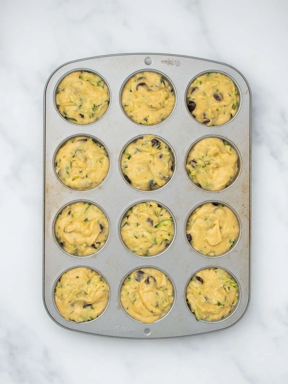 Standard metal 12-cup muffin tin with each well filled with light yellow raw muffin batter.