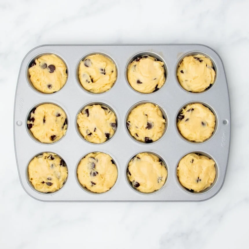 standard metal 12 cup muffin tin with raw yellow muffin batter with dark brown chocolate chips filling all 12 wells