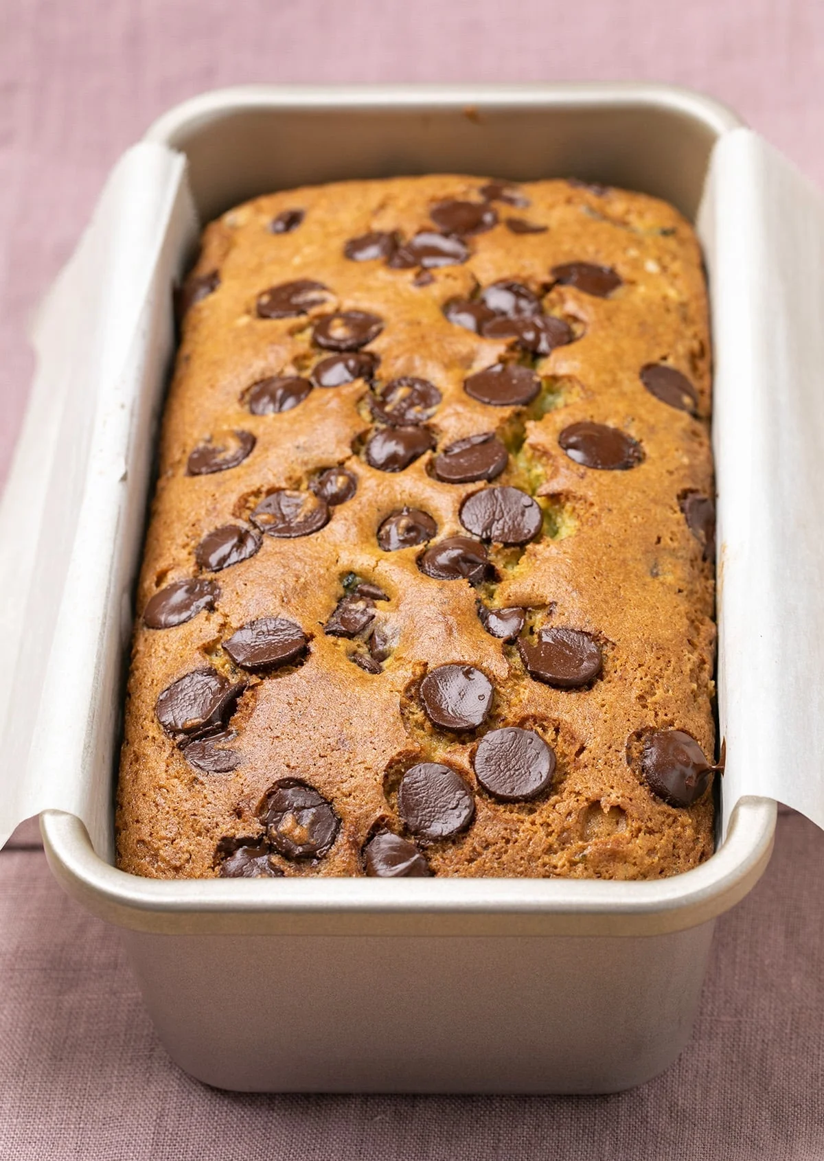 The bread with brown spots in nonstick loaf pan lined with white paper on purple cloth.