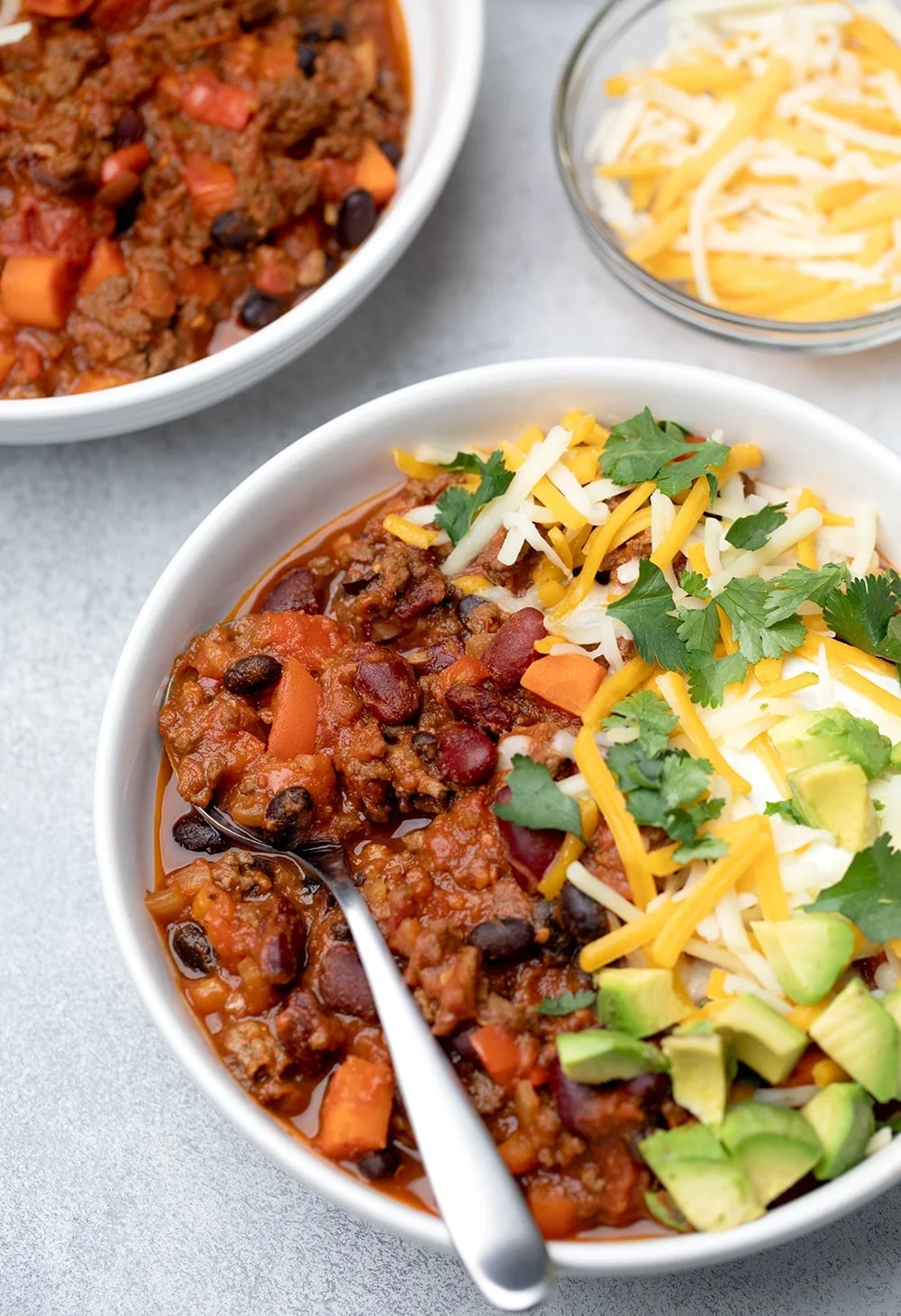 The chili served with green white and yellow toppings in bowl with spoon nearly overhead.