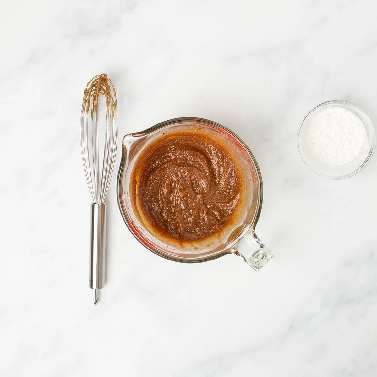 thick brown pasta in same glass measuring cup with wire whisk on marble table next to it