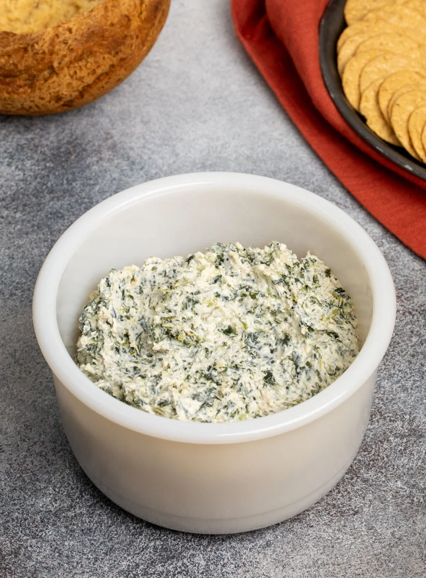 thick white and green flecked spinach dip in round deep white bowl on speckled gray surface with crackers on black plate and bread bowl in background