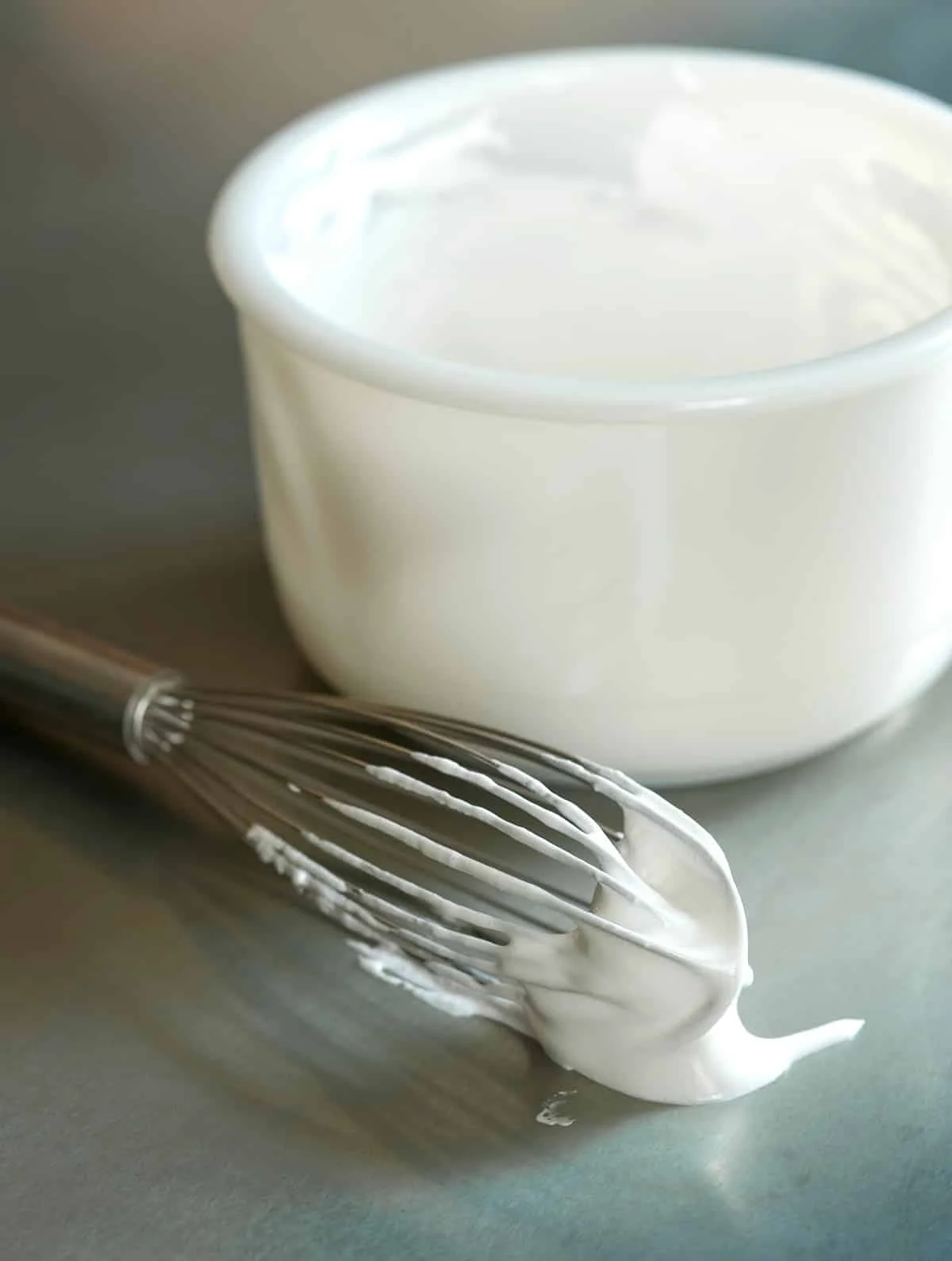 thick white marshmallow creme on wire whisk on metal table with white bowl in background