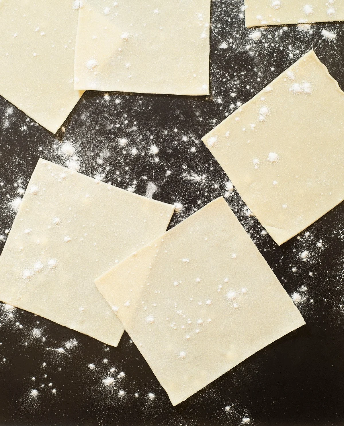 thin yellow squares of dough with white flour on black surface