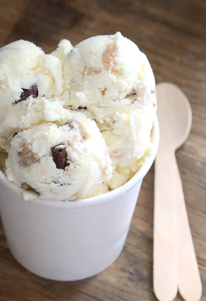 Three scoops of gf vanilla ice cream with gf cookie dough balls in a white pint container with wooden spoons