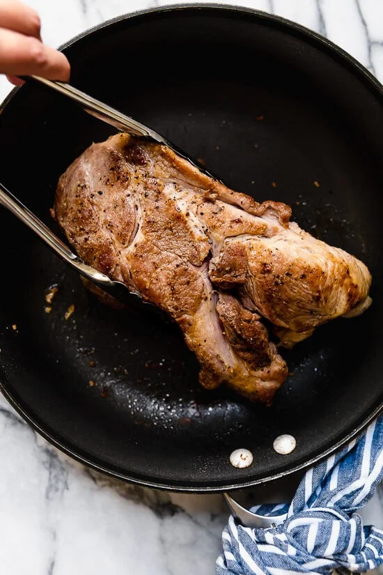 Tongs grabbing pork shoulder in skillet.