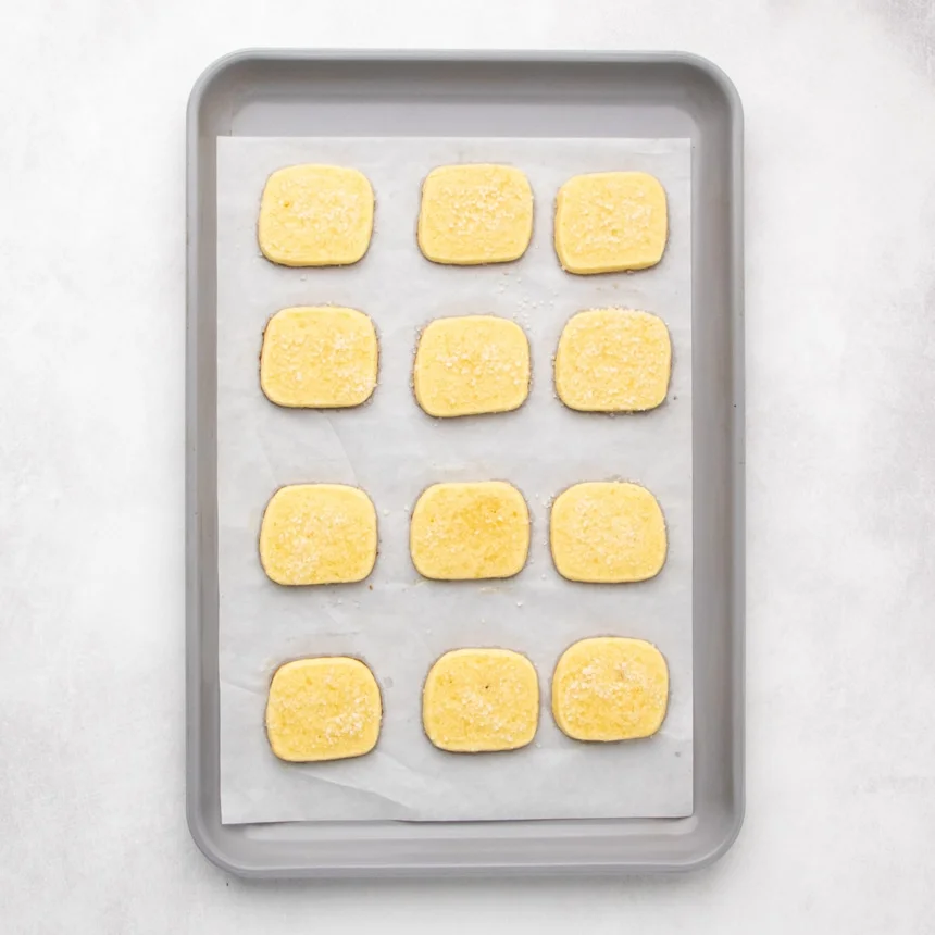 Twelve baked small rectangular yellow cookies with coarse white sugar grains baked into the top, arranged in rows on a parchment-lined baking sheet.