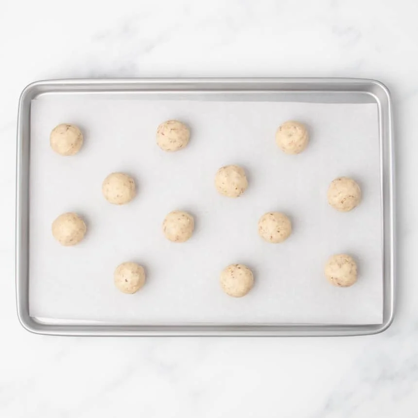 Twelve rounded raw balls of light tan cookie dough arranged evenly on a parchment-lined baking sheet.