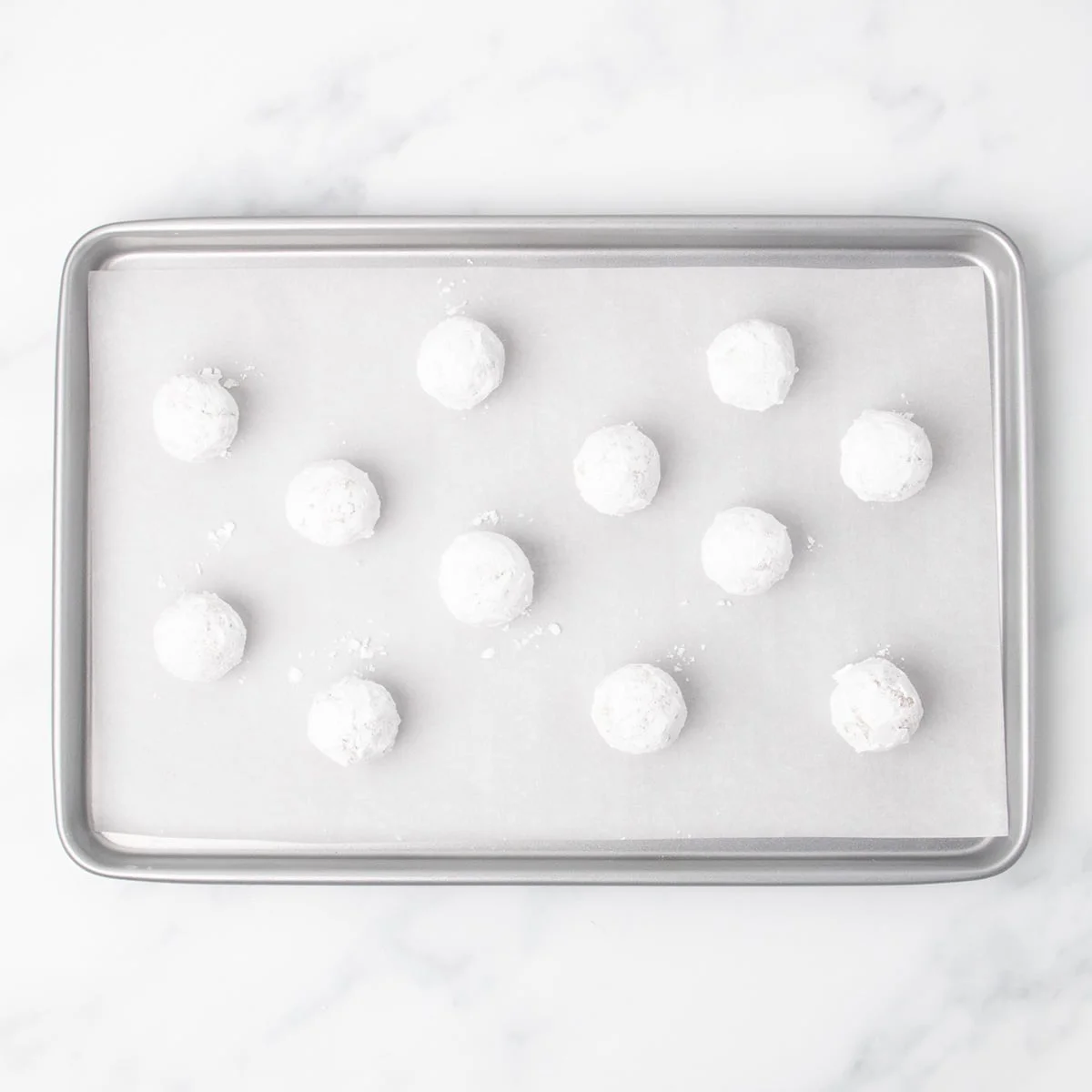 Twelve white appearing raw sugar-coated dough balls arranged evenly on a parchment-lined baking sheet.
