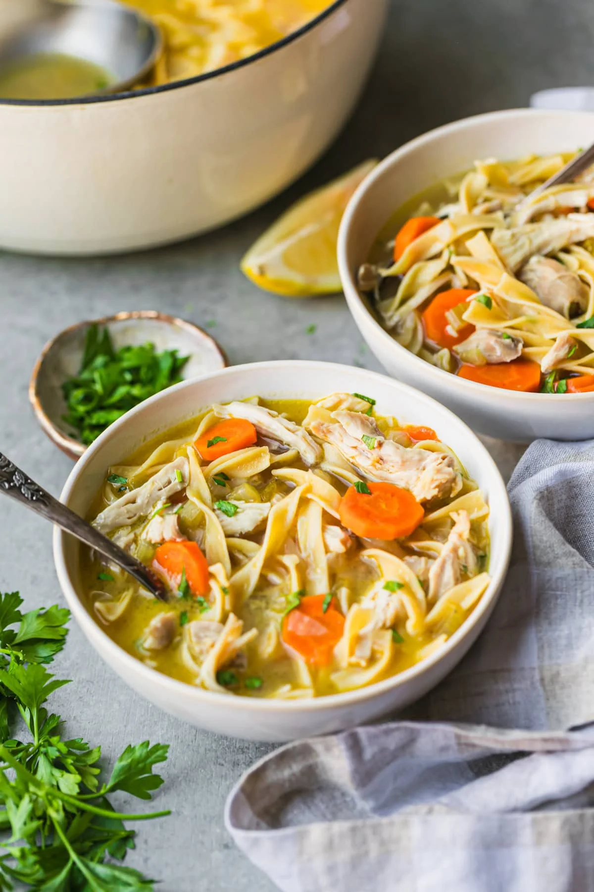Two bowls of chicken noodle soup next to chopped fresh parsley and a lemon wedge.