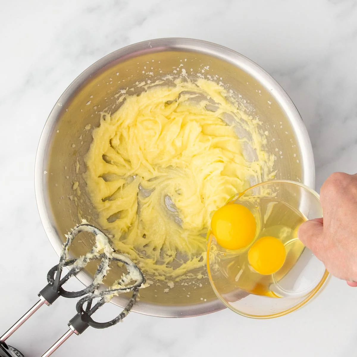 Two eggs being added to a pale yellow creamed butter and single egg mixture with electric beaters