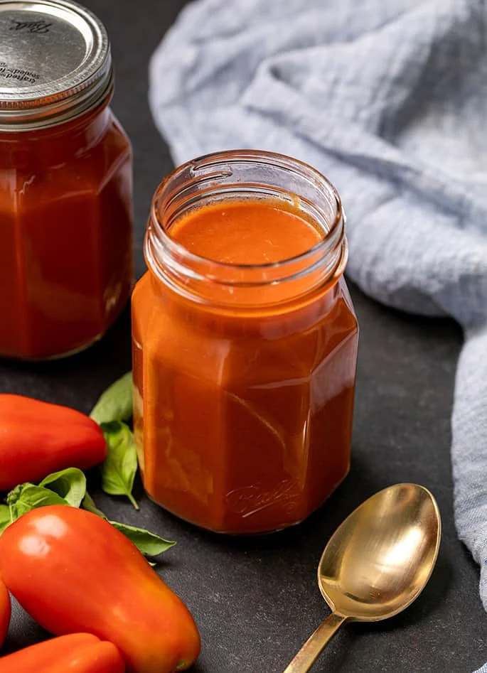 Two glass jars with red gluten free enchilada sauce, one with a lid, blue cloth, gold soup spoon, plum tomatoes and basil leaves