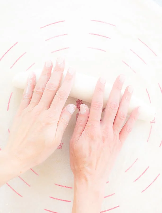 Two hands shaping a loaf of raw french bread dough on a canvas cloth with red lines