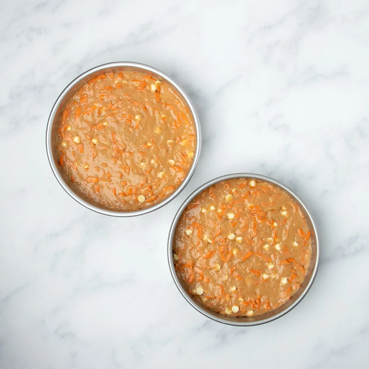 Two round 8-inch metal cake pans with wet raw gluten free carrot cake batter with visible carrot shreds and white chocolate chips.