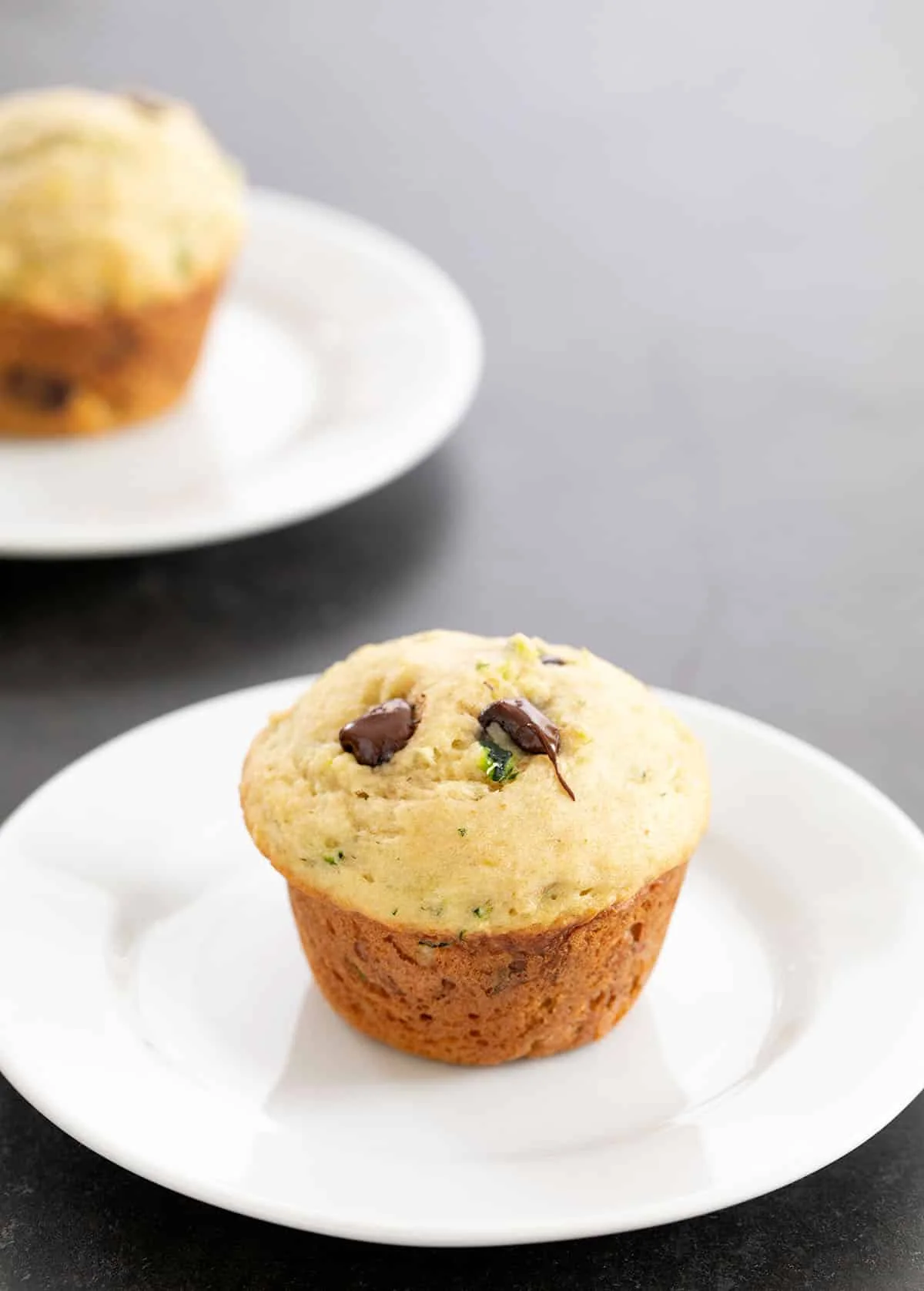 Two zucchini muffins each on a small white plate on a gray surface.