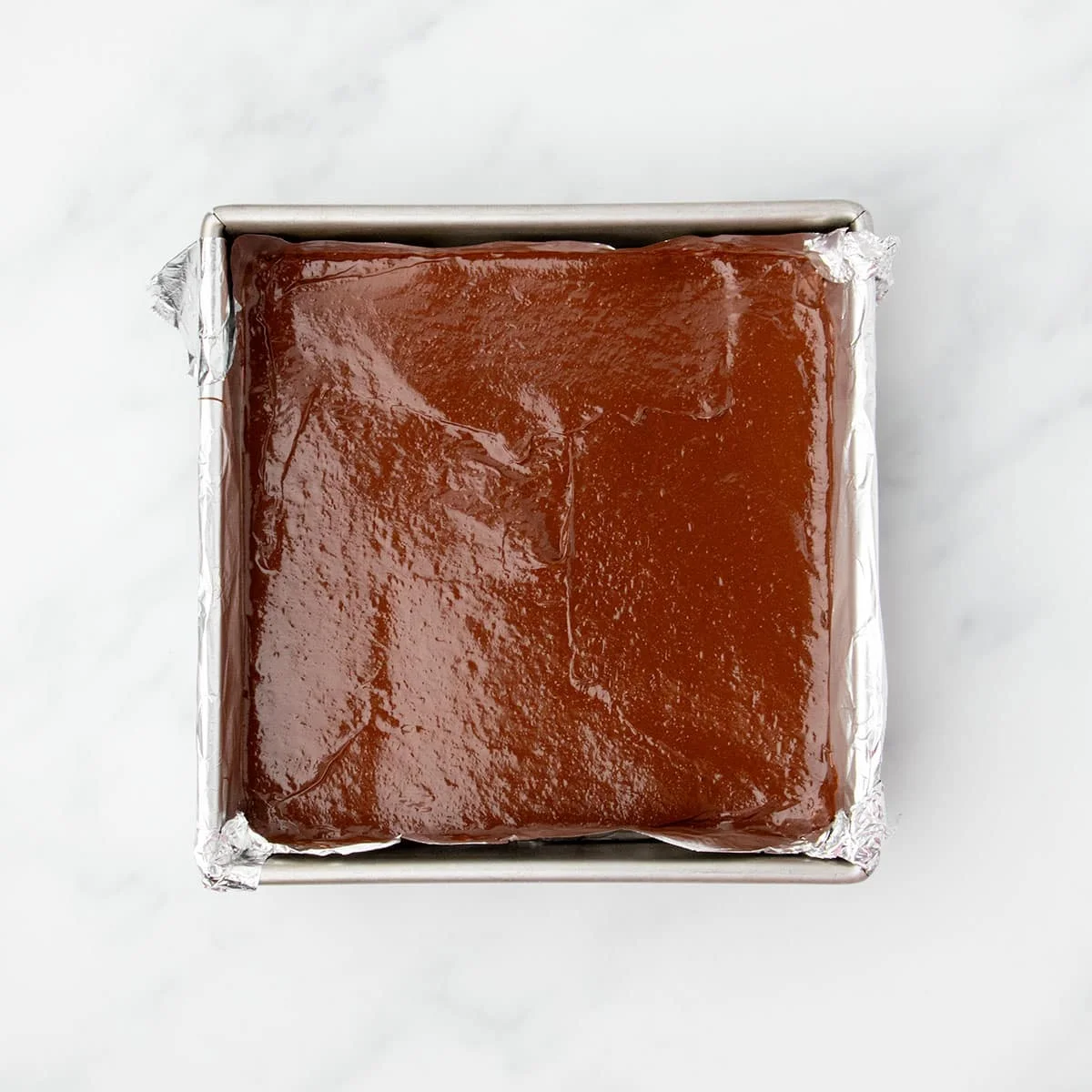 Unbaked brownie batter spread evenly in a foil-lined square metal pan