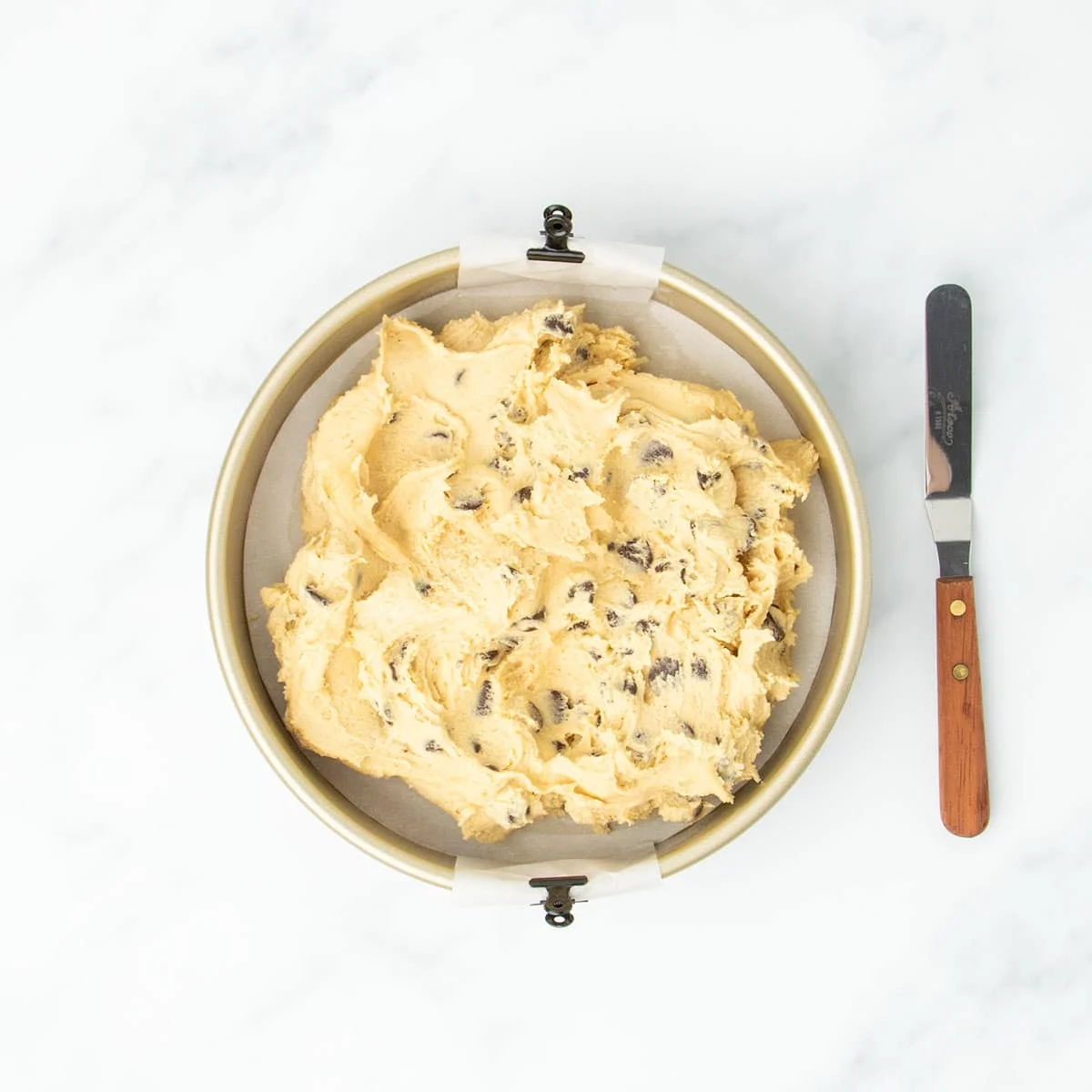 Unbaked raw chocolate chip cookie dough spread roughly in a gold colored round baking pan lined with parchment