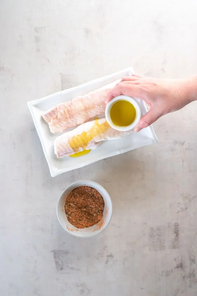Uncooked cod on a white plate, pouring oil on the fish.