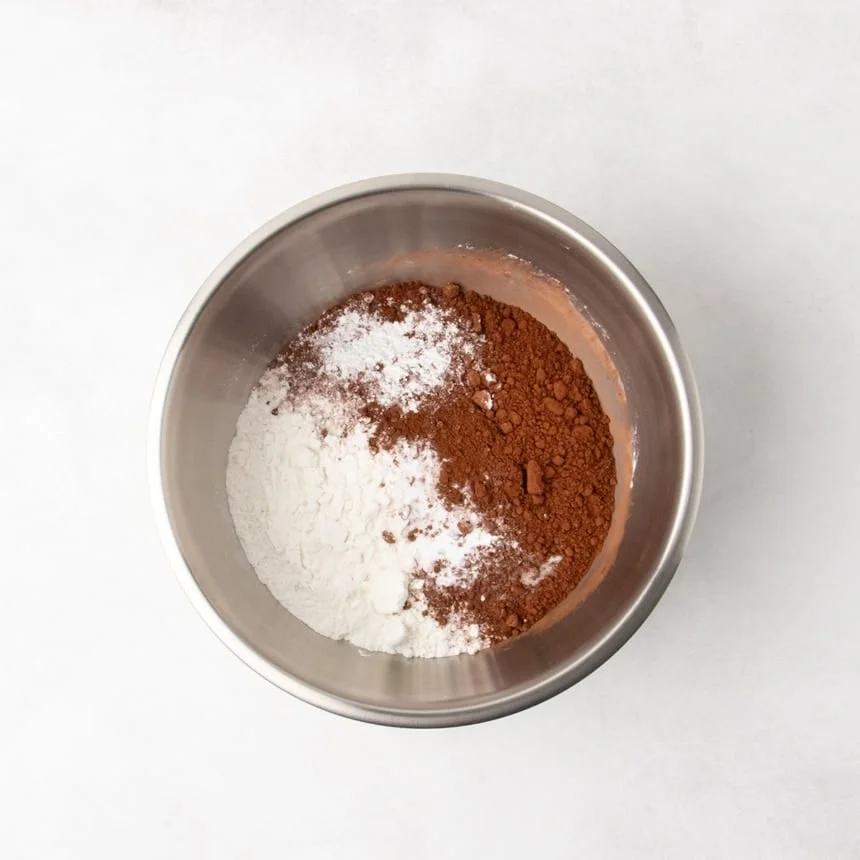Unwhisked ingredients in a stainless steel mixing bowl, with white powder on one side and brown powder on the other.