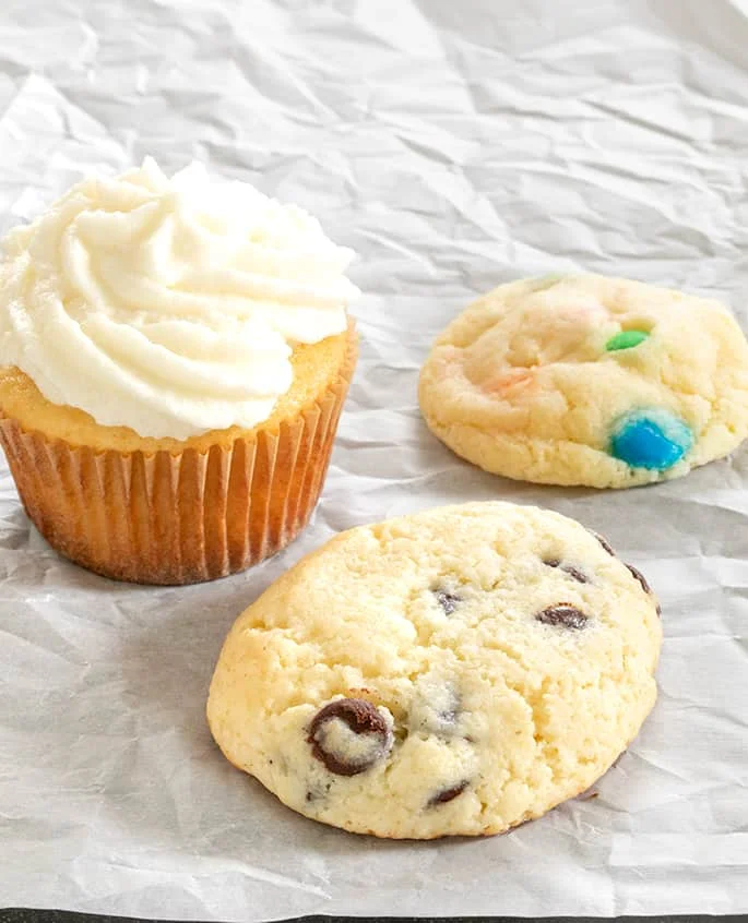 Vanilla frosted cupcake, cookie with M&Ms and a chocolate chip cookie on white surface 