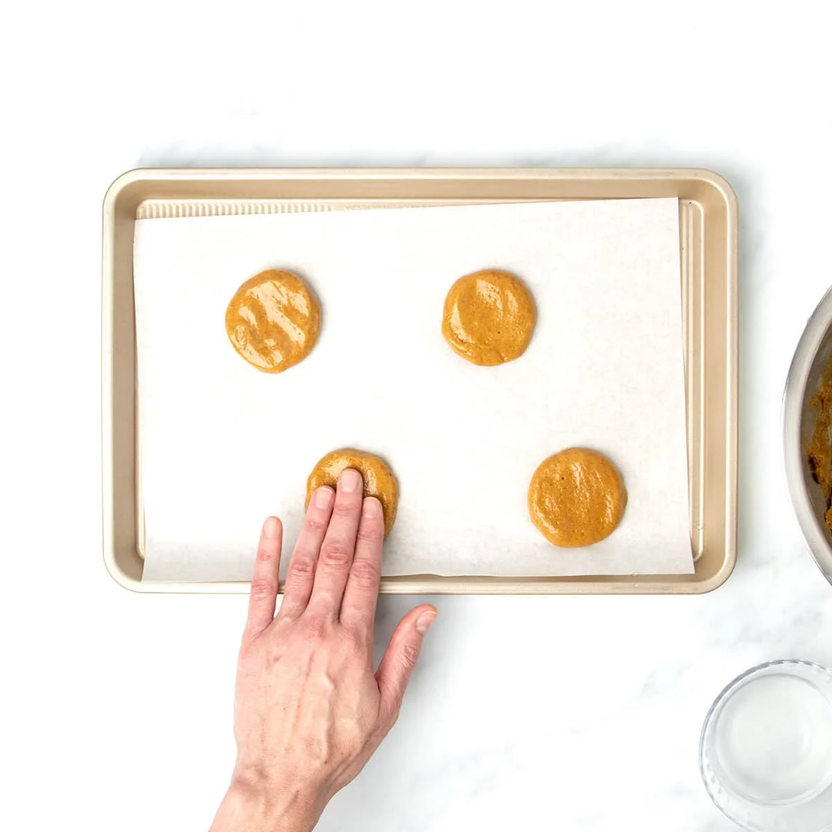 wet fingers pressing down on light brown round of cookie dough into a disk on white paper on gold baking tray