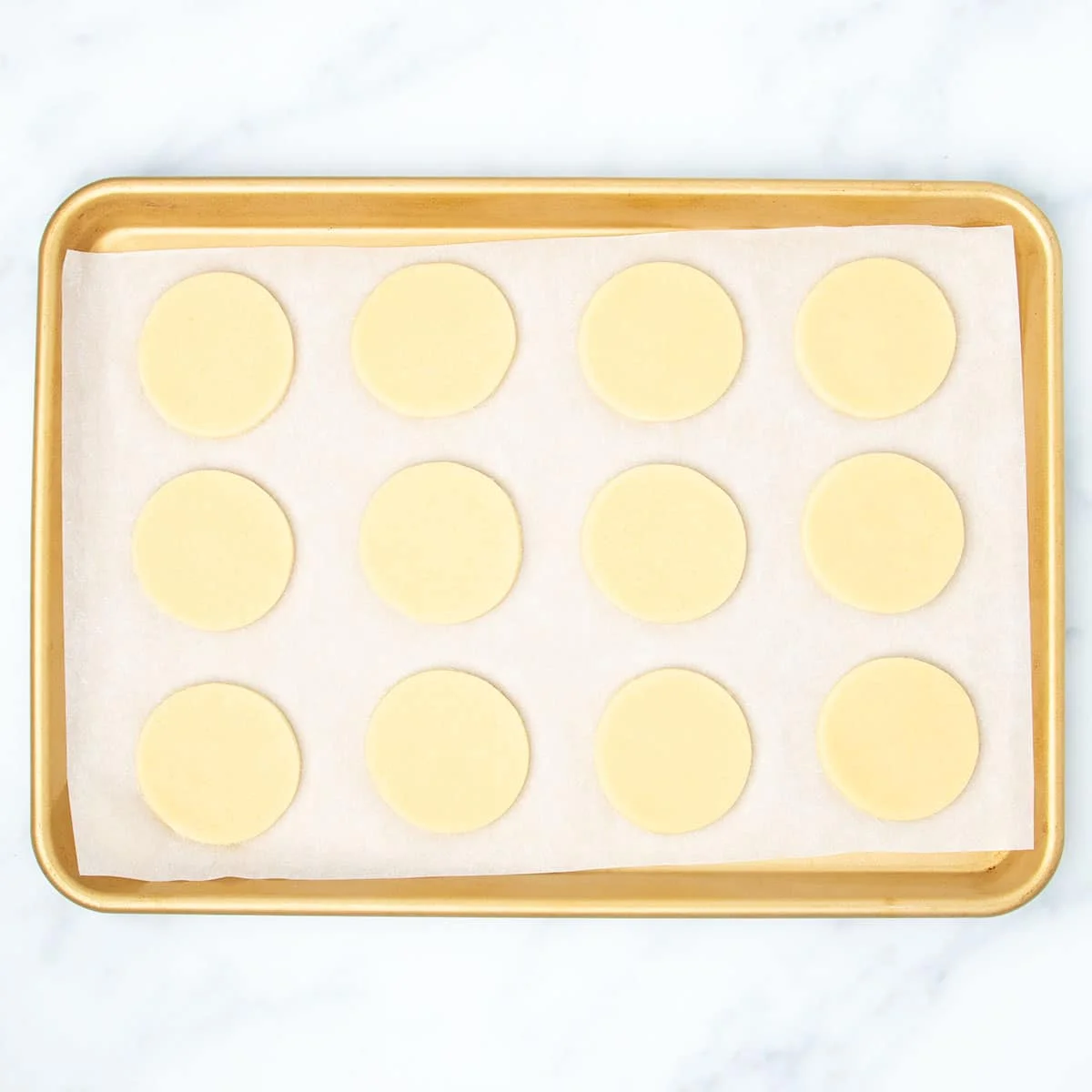White paper lined gold rectangular baking sheet with 12 raw rounds of sugar cookie dough.