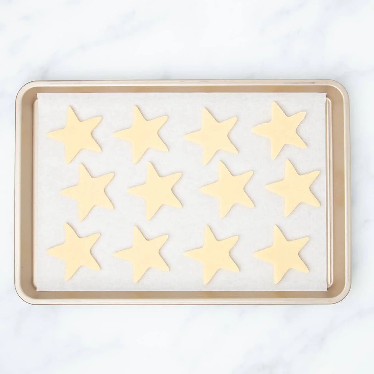 White paper lined gold rectangular baking sheet with 12 raw stars of sugar cookie dough.