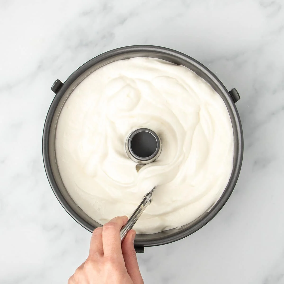white soft angel food cake batter in black metal tube pan with hand holding knife in mixture