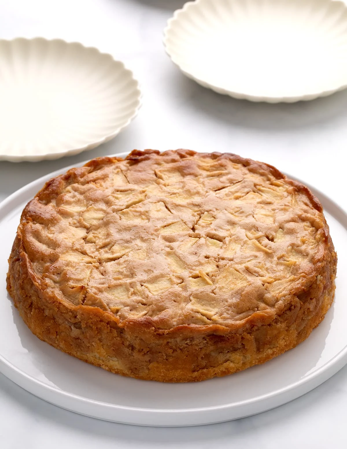 Whole round golden brown cake with visible cooked apple pieces on a white platter, small white cake plates in the background.
