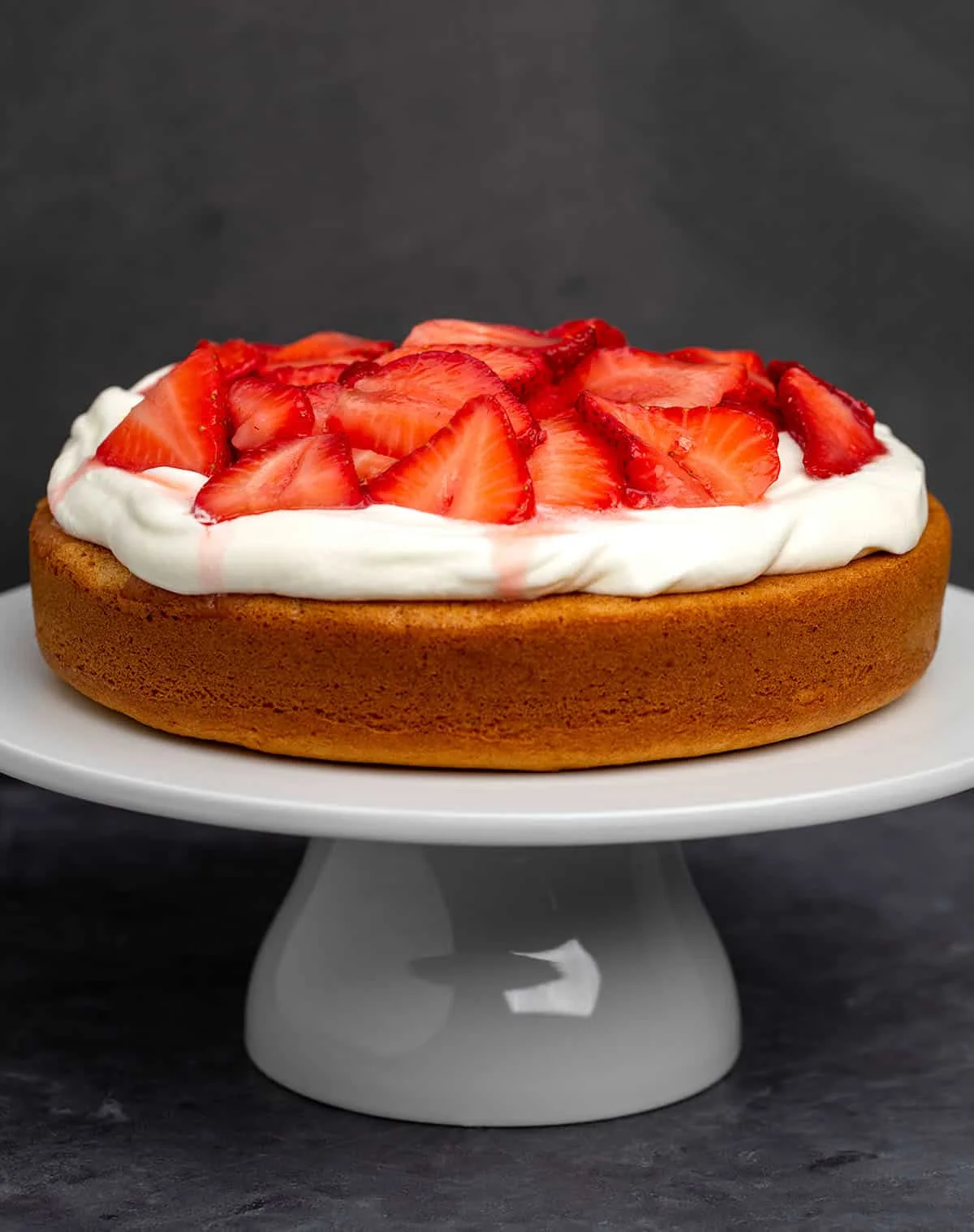 whole single layer vanilla cake with light brown exterior with a thick layer of white whipped cream and sliced strawberries with running red juice on white cake plate on black marbled surface