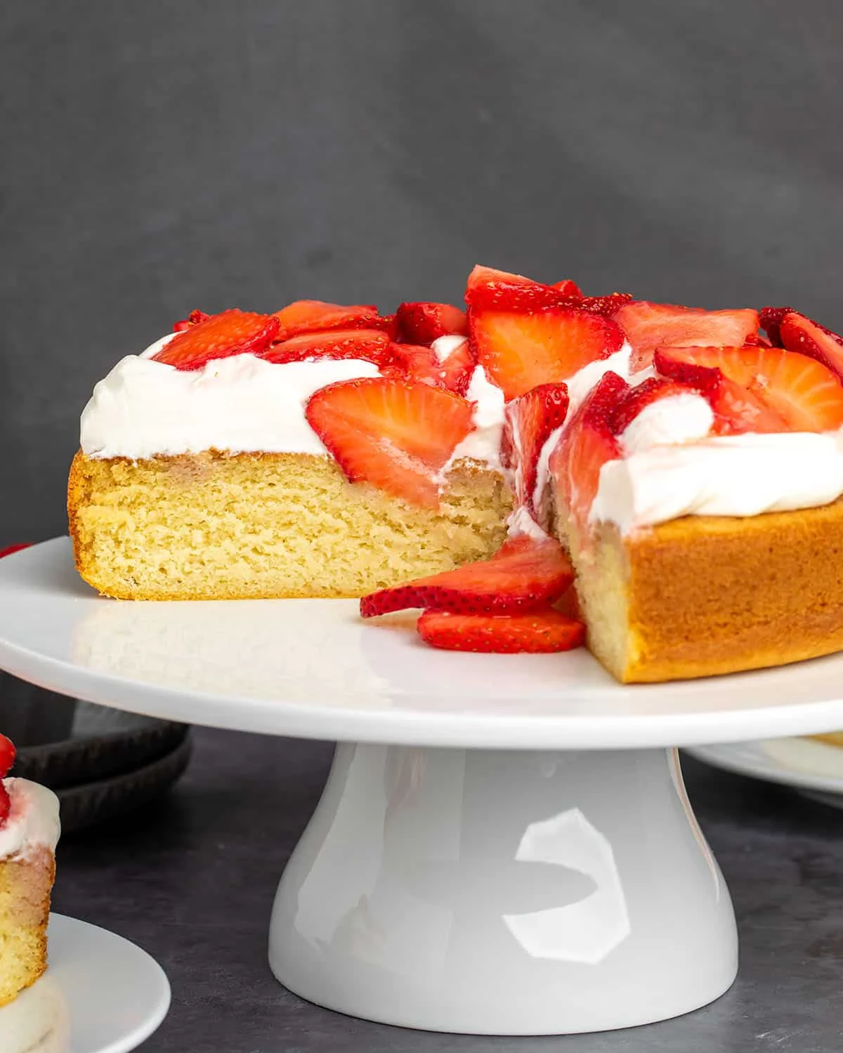 yellow cake with 2 slices missing with thick layer of white whipped cream on top and sliced red strawberries on a white cake plate with a black background